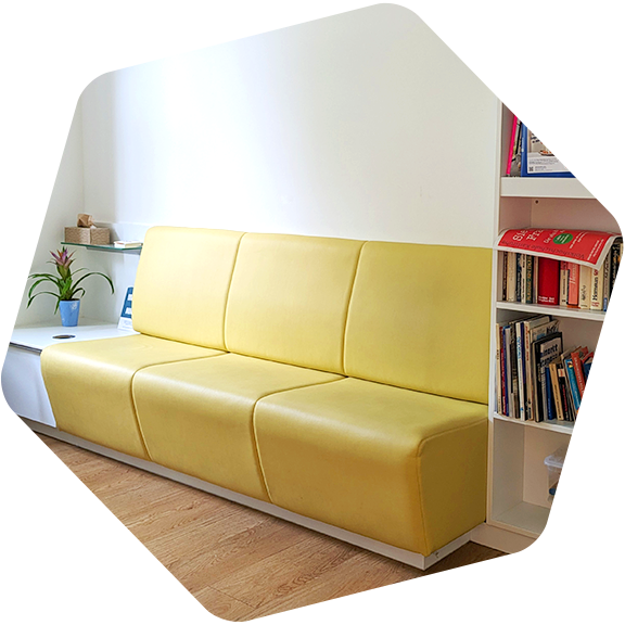 an der Wand entlang, ein Regal mit Büchern, eine gelbe Coach, ein kleiner Tisch mit Topfpflanze